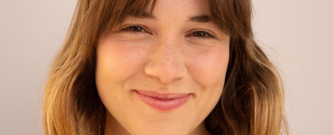 A person with shoulder-length brown hair smiles warmly at the camera against a neutral background.