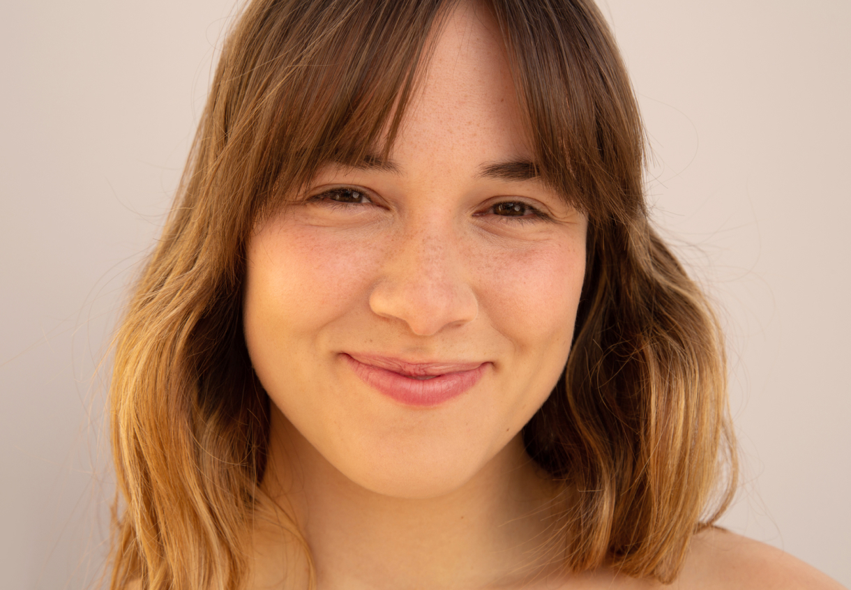 A person with shoulder-length brown hair smiles warmly at the camera against a neutral background.