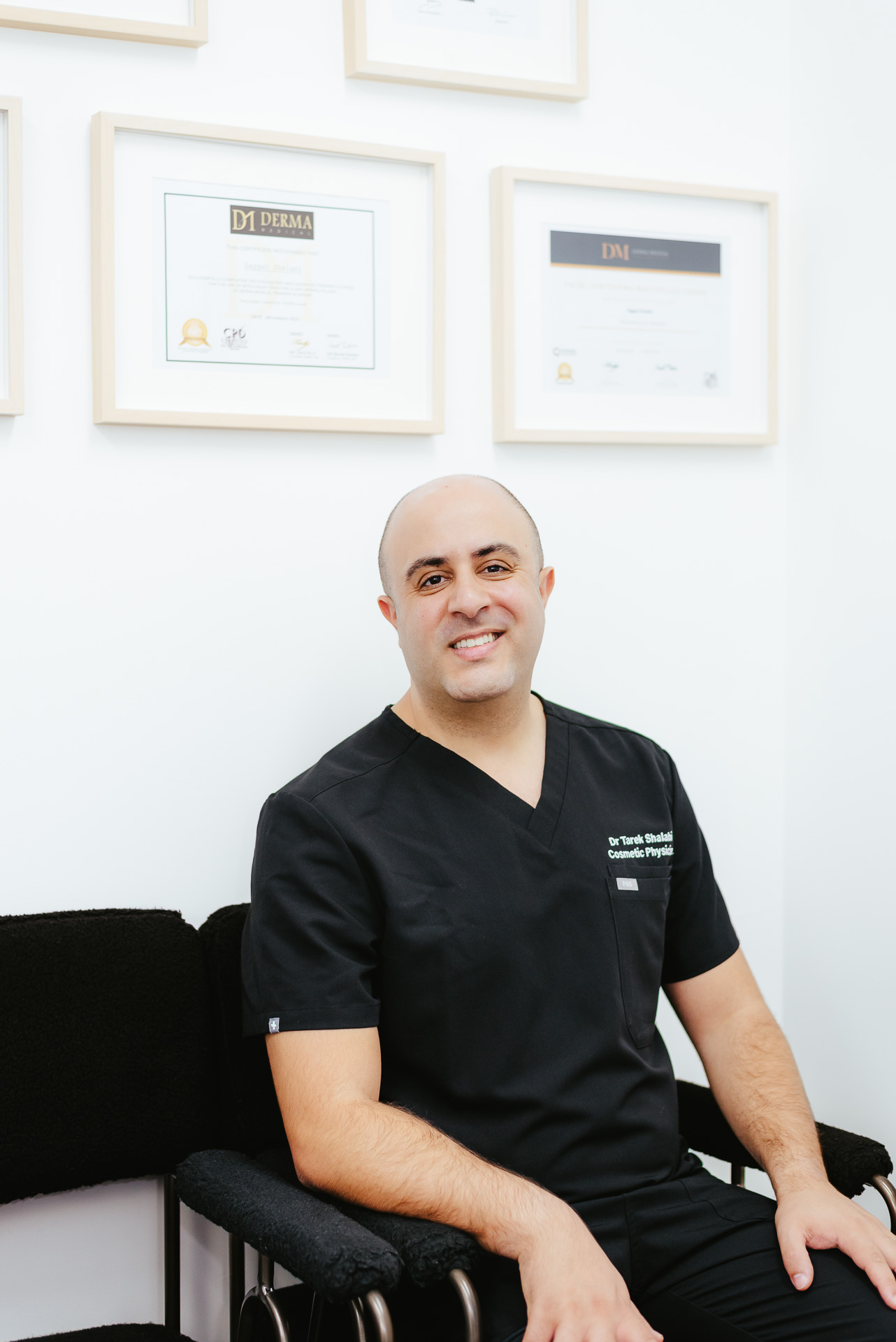 A person in black medical scrubs is seated on a chair in an office. They are smiling and have a clean-shaven head. The background features framed certificates on a white wall.