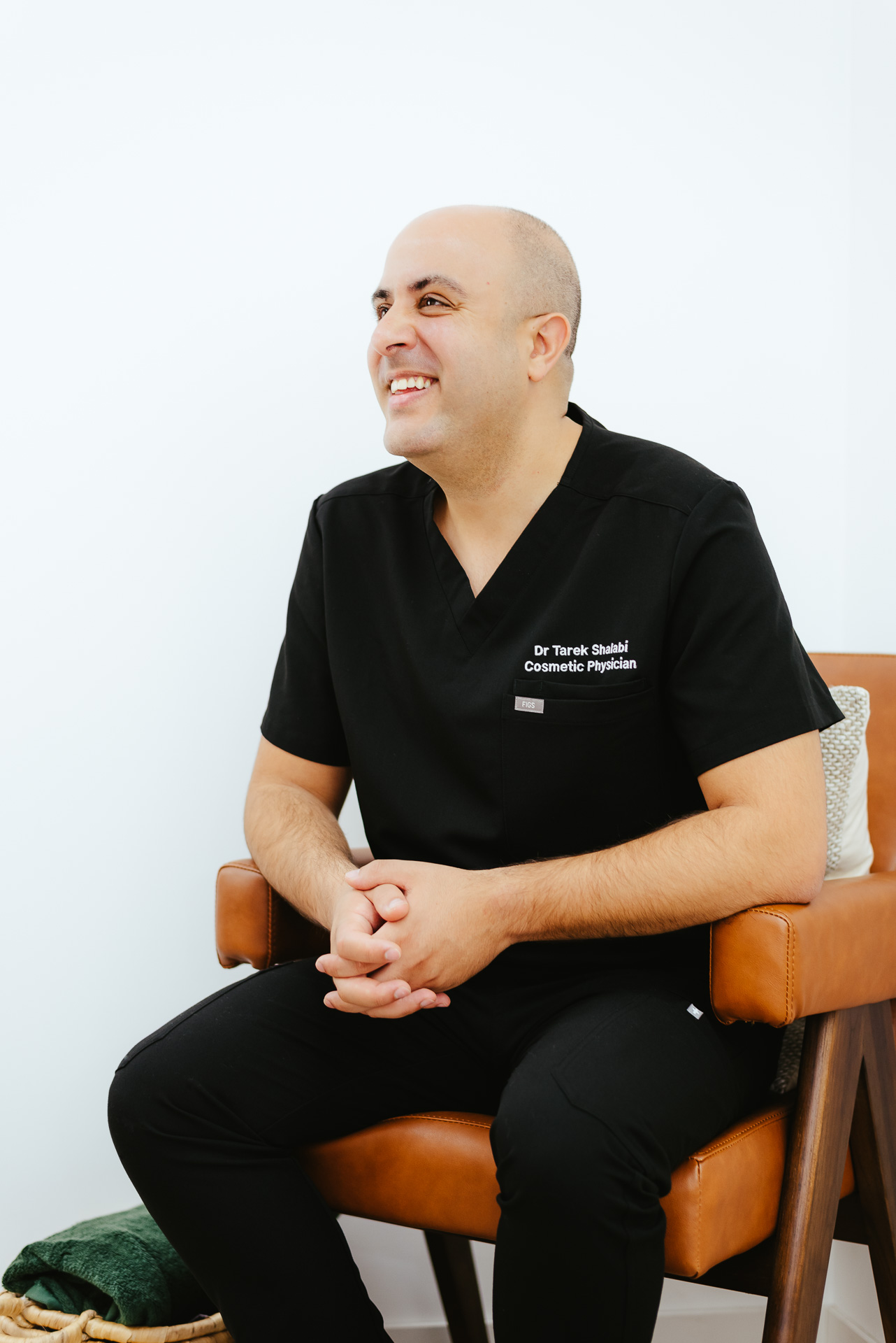 A man with a shaved head, wearing black scrubs that say "Dr. Tarek Shaief, cosmetic physician," sits on a brown chair. He is smiling and looking upwards. A green cushion and a wicker basket are nearby.