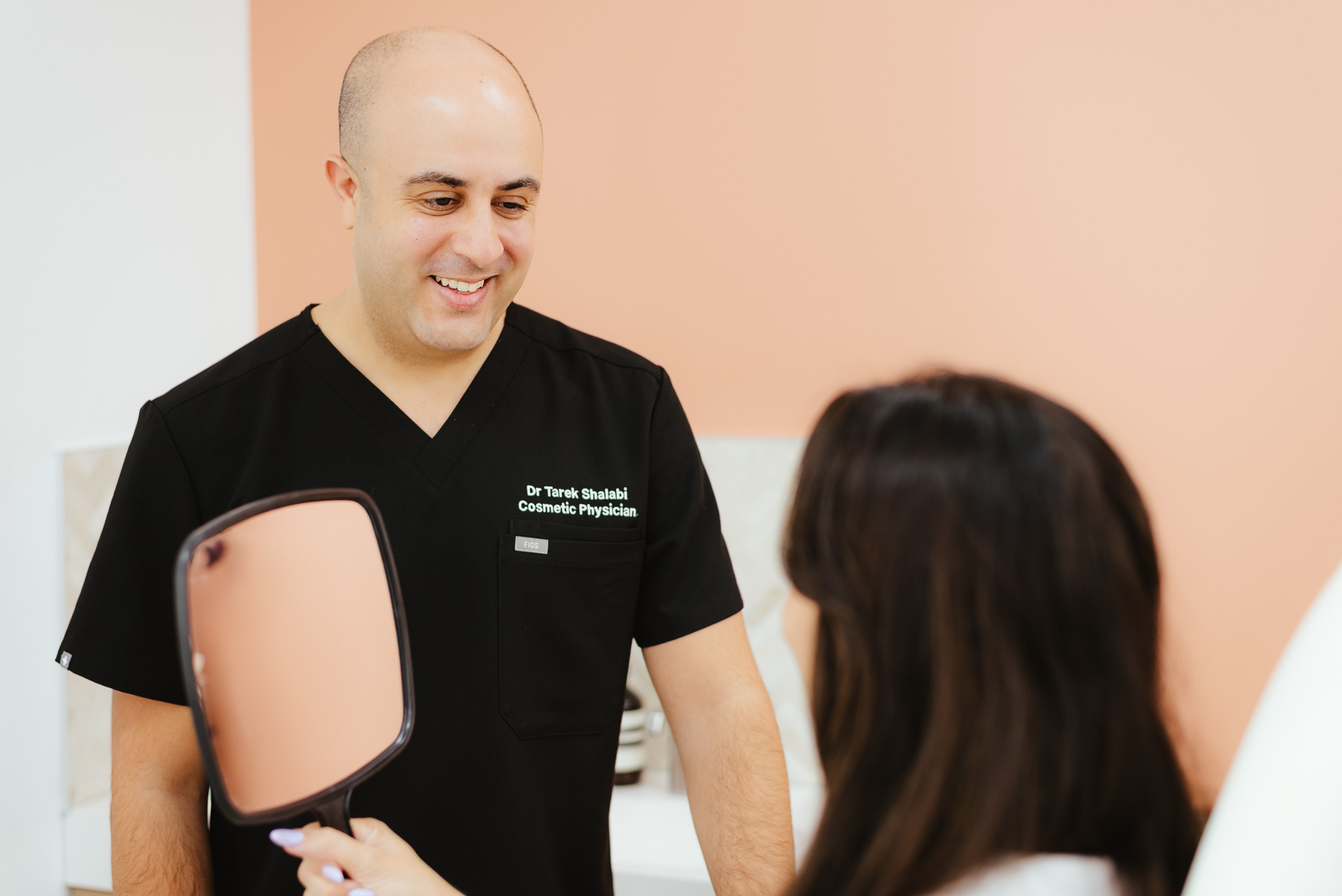 A cosmetic physician in a black uniform smiles at a person holding a handheld mirror in a clinic setting with peach-colored walls.