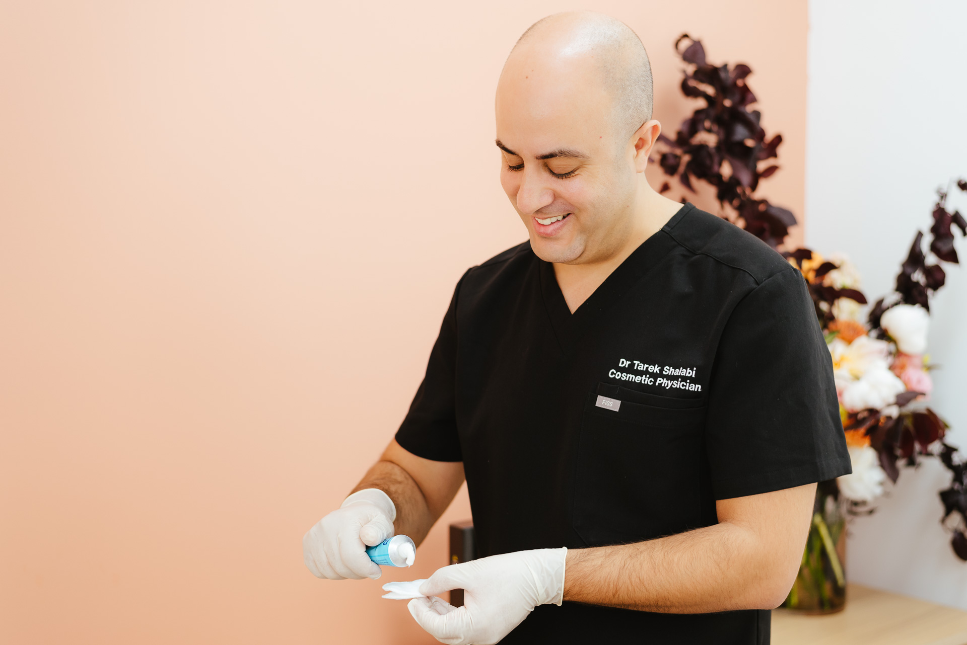 A smiling person in black scrubs and white gloves prepares a medical product on a cotton pad, against a soft peach and white background with floral arrangements.