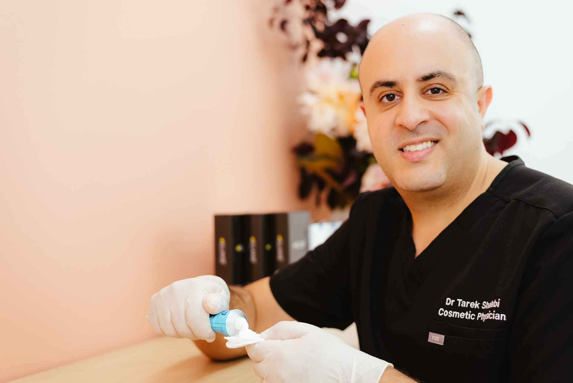 A bald man in a black medical uniform is holding a small container and applying cream onto a pad. He is wearing gloves and smiling, standing in a room with a peach wall and floral decor. Several product containers are on the counter.