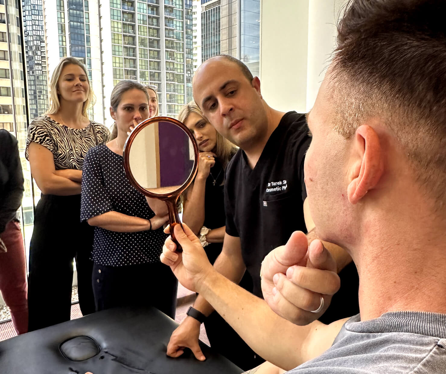 A man sits holding a mirror, examining his face while a medical professional explains something, touching his jawline. A group of four people stands nearby, attentively observing the demonstration. The background shows tall city buildings.