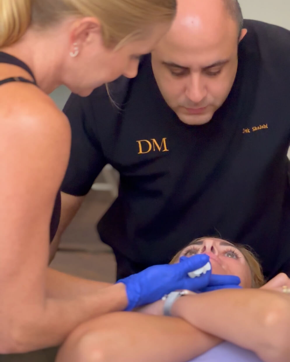 A person lying down receives a cosmetic treatment from a professional wearing gloves, assisted by another person wearing a black shirt with initials "DM." The setting appears clinical, focused on facial care.