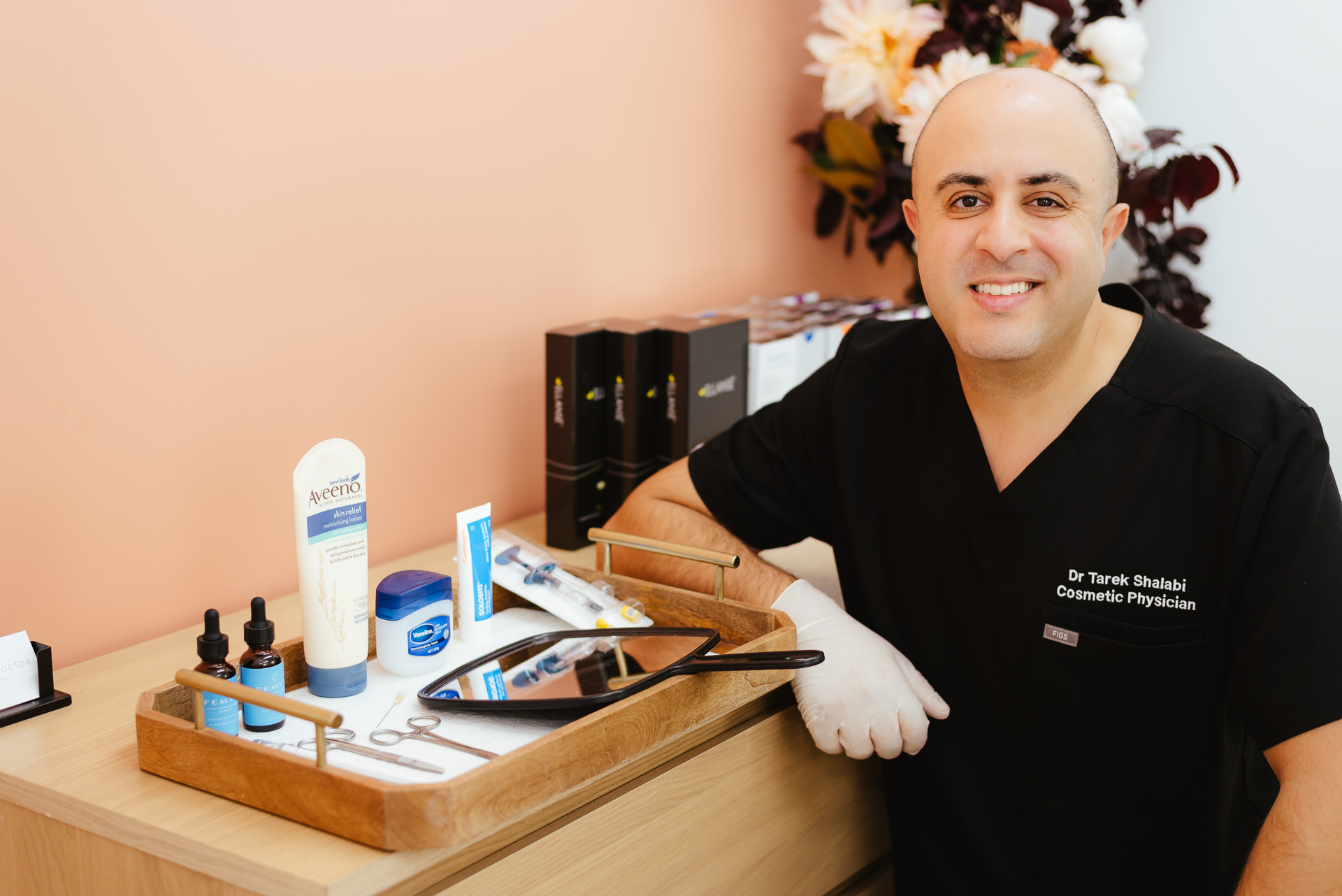 A physician in a black uniform stands next to a tray of skincare products and tools, smiling at the camera. Behind him, there is a peach-colored wall with white and dark flowers in a vase.