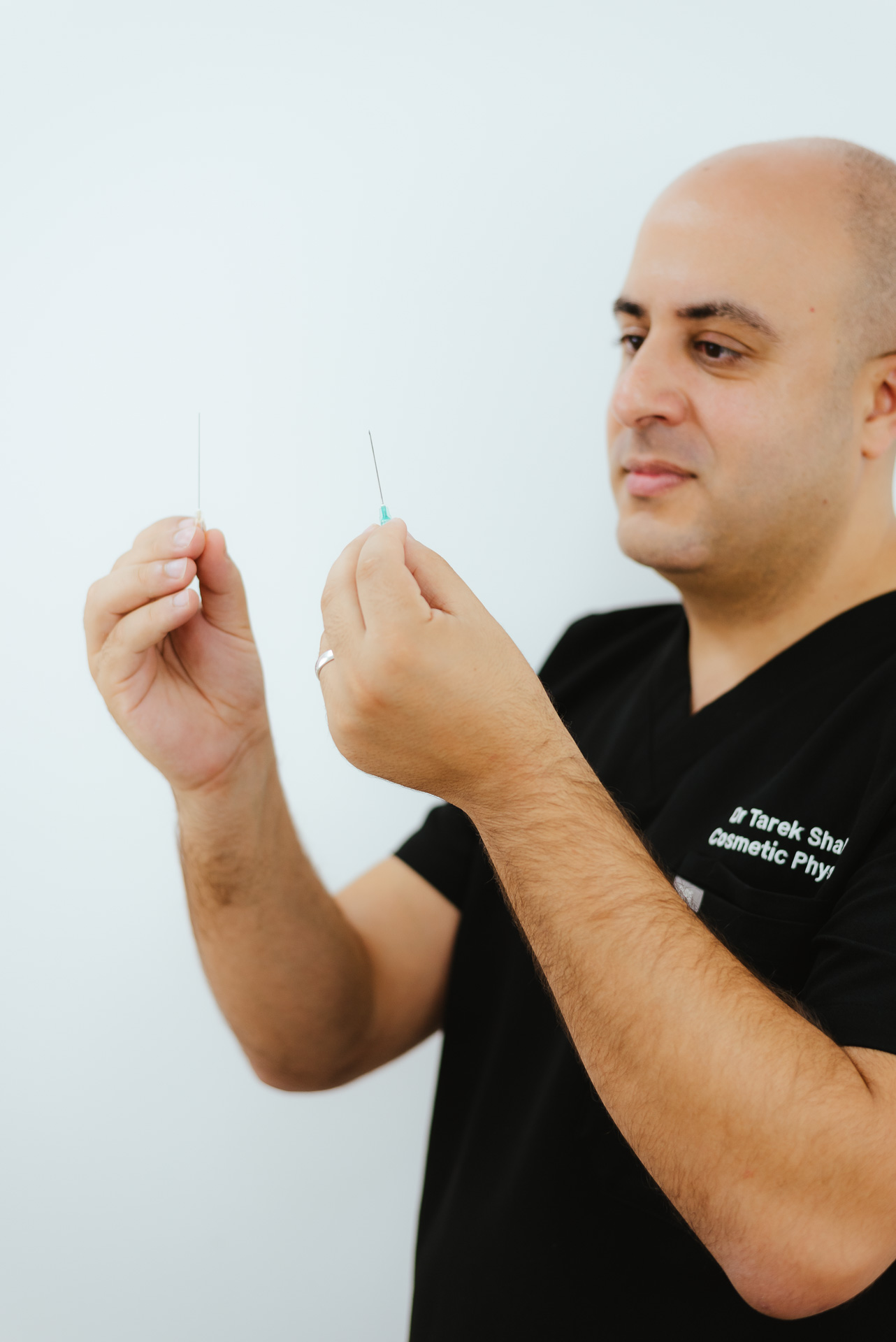 A person in a black scrub top holds two needles, examining them closely, against a plain background.