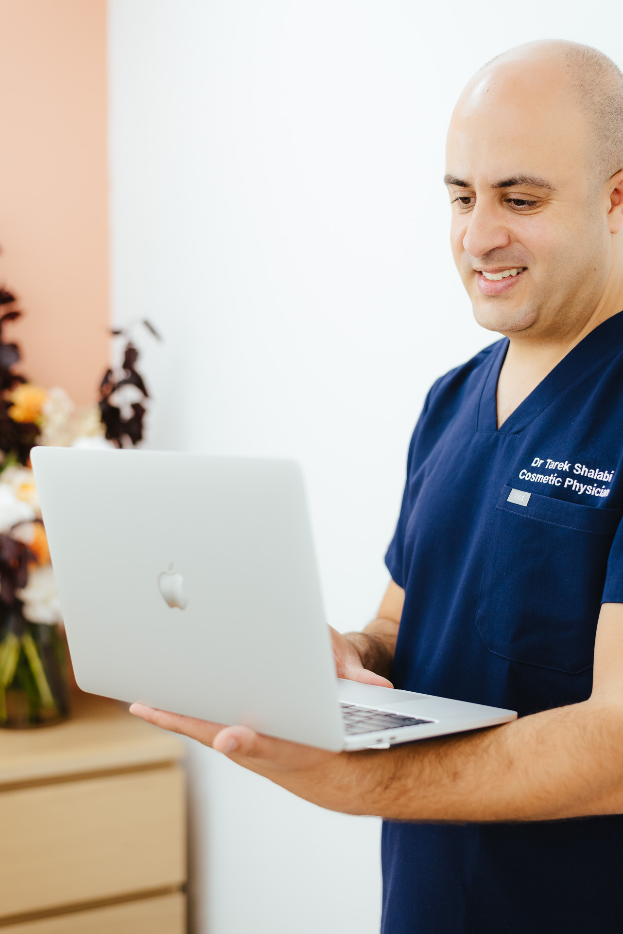 A person in navy scrubs is smiling while looking at a laptop. The background features a light-colored wall and a bouquet of flowers.