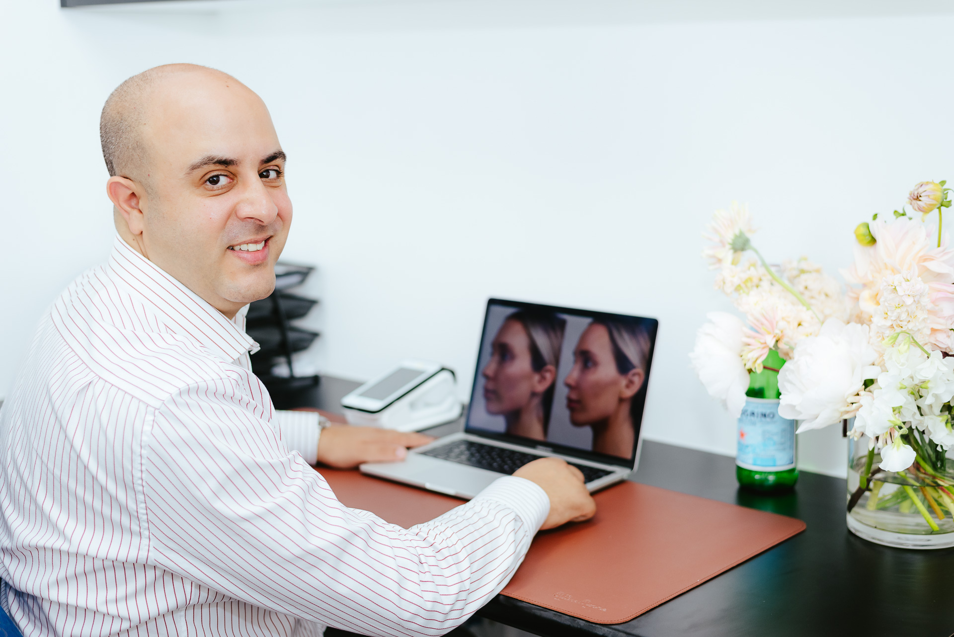 A person sitting at a desk smiles at the camera. They are working on a laptop showing side-by-side images of a face. The desk has a phone, a stack of trays, and a vase with flowers.