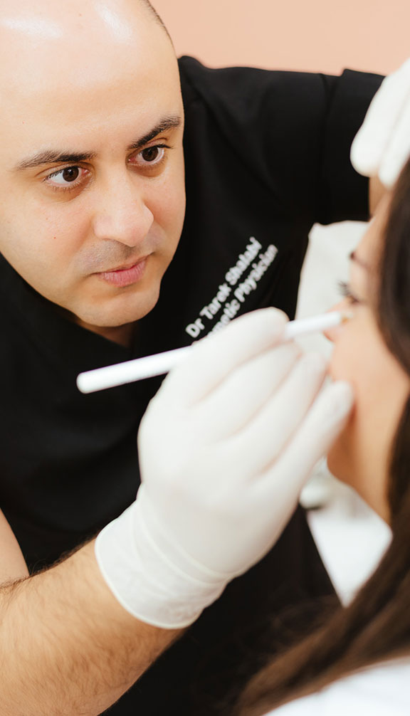 A person wearing gloves uses a white pencil tool near a woman's face in a clinical setting. The individual appears focused and professional. Both are engaged in a cosmetic or medical procedure.