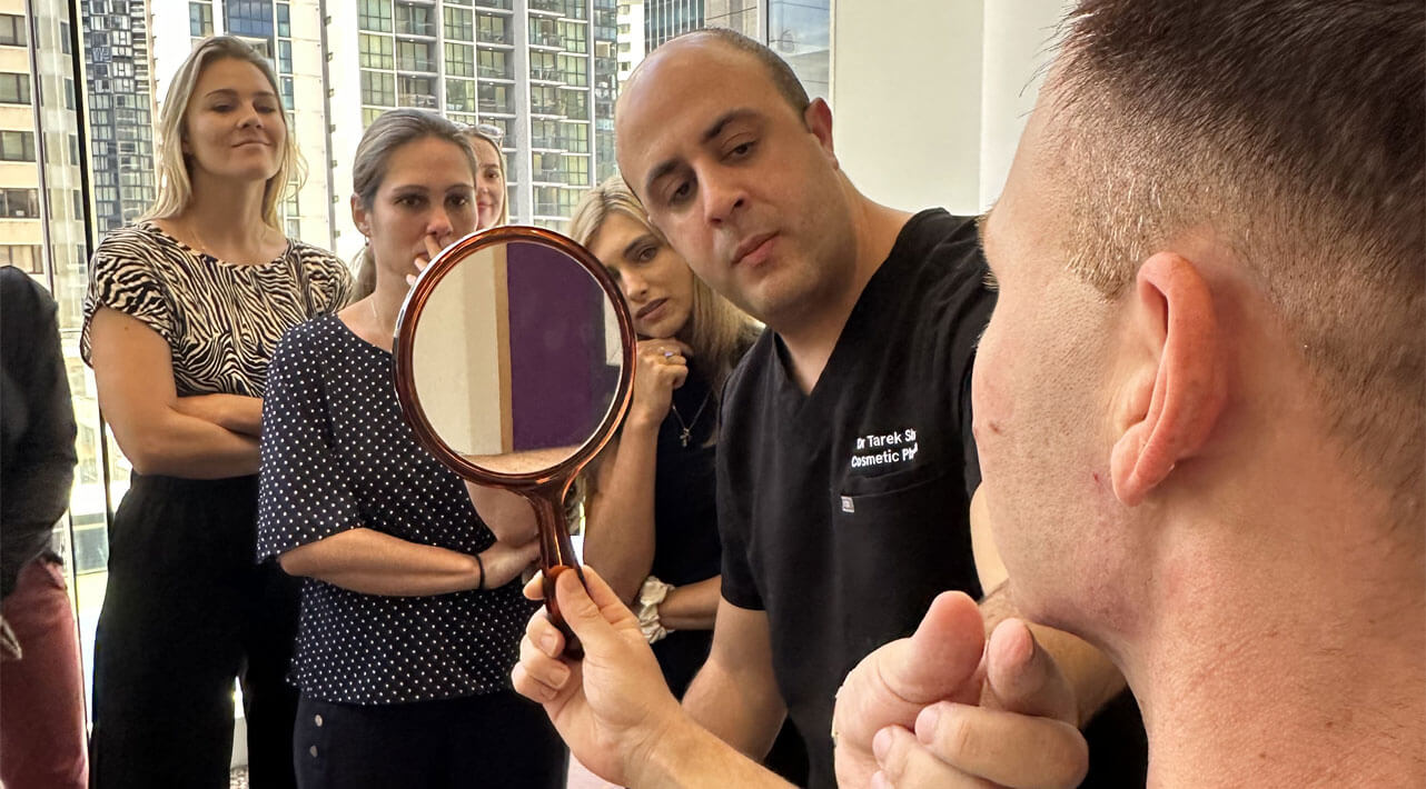 A group of people attentively watching a man holding a circular mirror as another man, wearing dark scrubs, points to his face. They are in a room with large windows showing tall buildings outside.