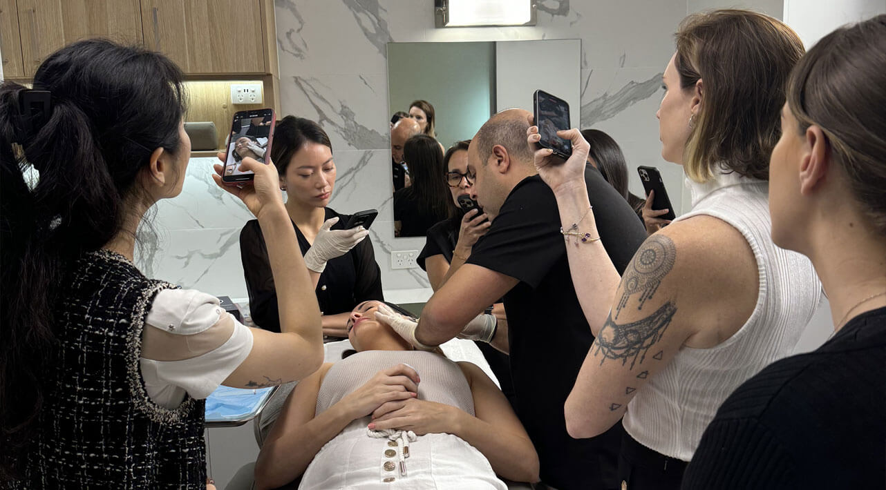 A group of people, some holding phones, surround a person receiving a cosmetic procedure in a clinic. The room has marble walls and wood accents, and a mirror reflects the scene.