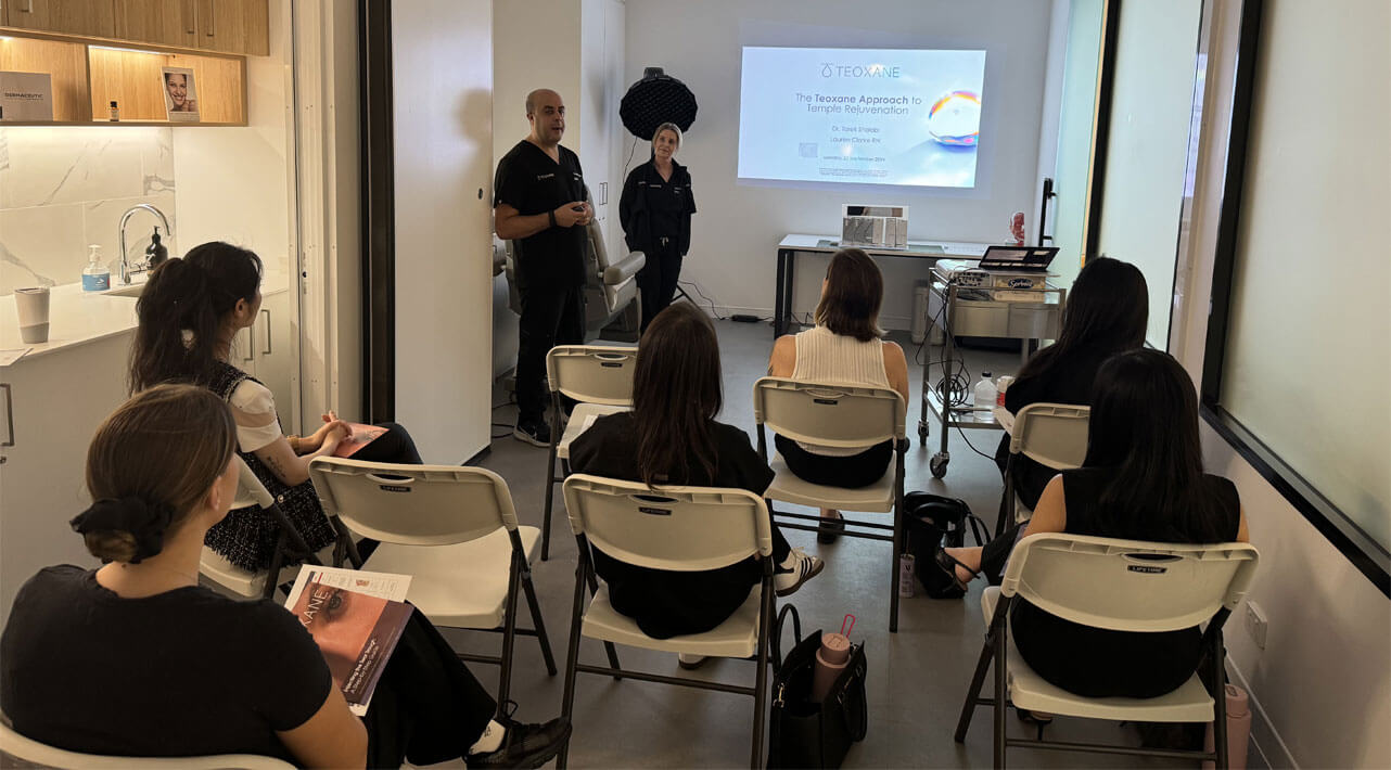 A man presents at the front of a small classroom, with a woman standing nearby. Several attendees sit on folding chairs, facing a projector screen displaying a presentation titled "The Research Approach to Professional Education.