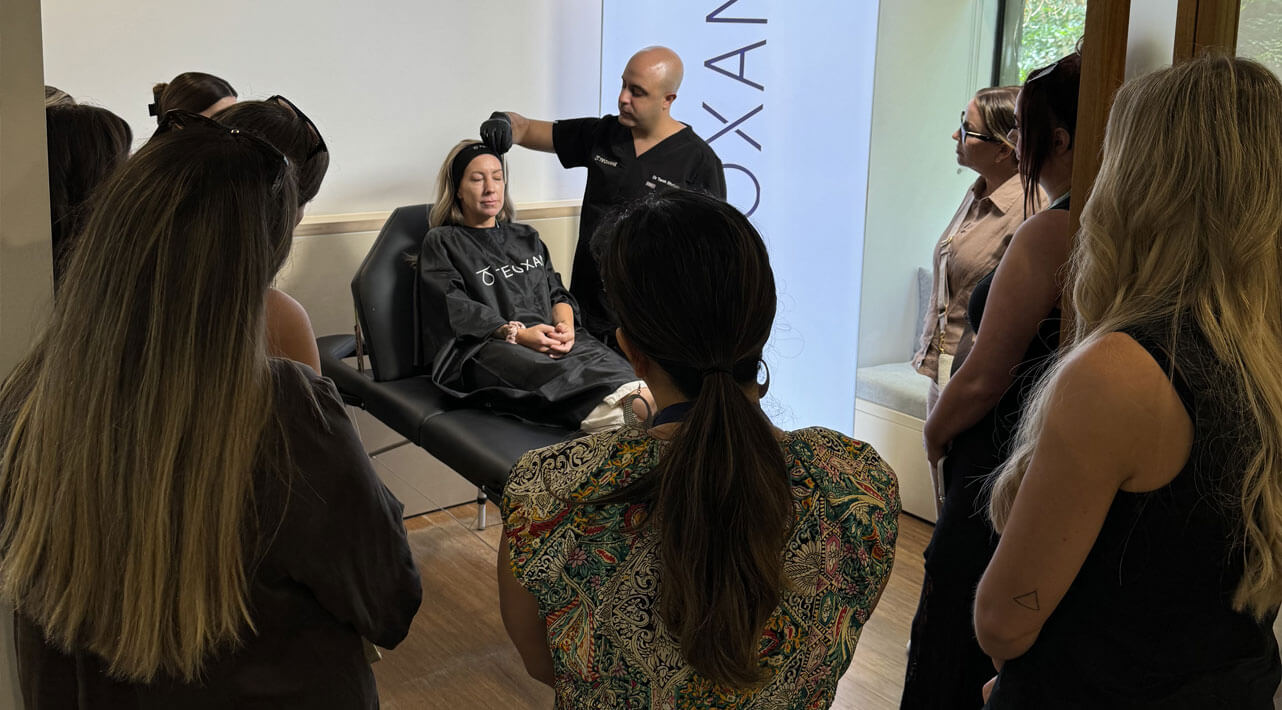 A group of people surrounds a woman lying on a reclined chair while a man, possibly an instructor, gestures and speaks. The setting resembles a medical or educational demonstration.